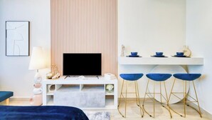 Una camera d'albergo moderna con una televisione a schermo piatto, una scrivania bianca con sedie azzurre, un comodino con una lampada e un quadro appeso al muro.