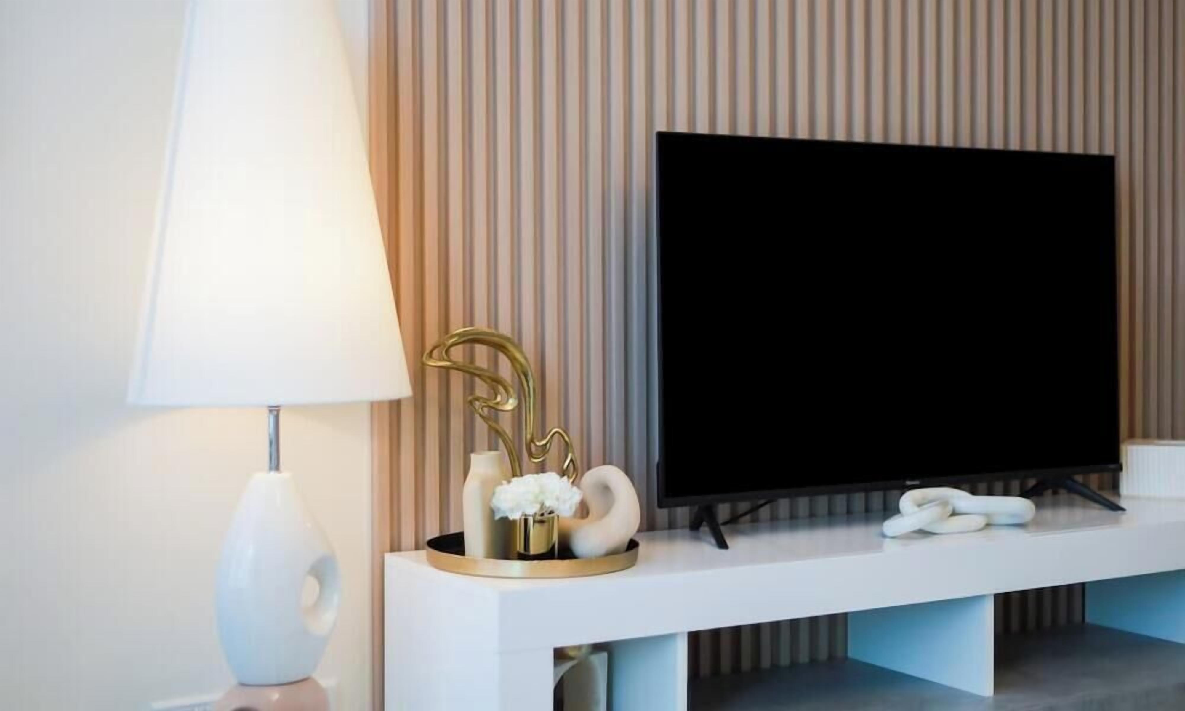 Un salon moderne avec un téléviseur à écran plat posé sur une console blanche, un lampadaire blanc et des objets décoratifs, notamment un vase doré et un objet en forme de cygne.