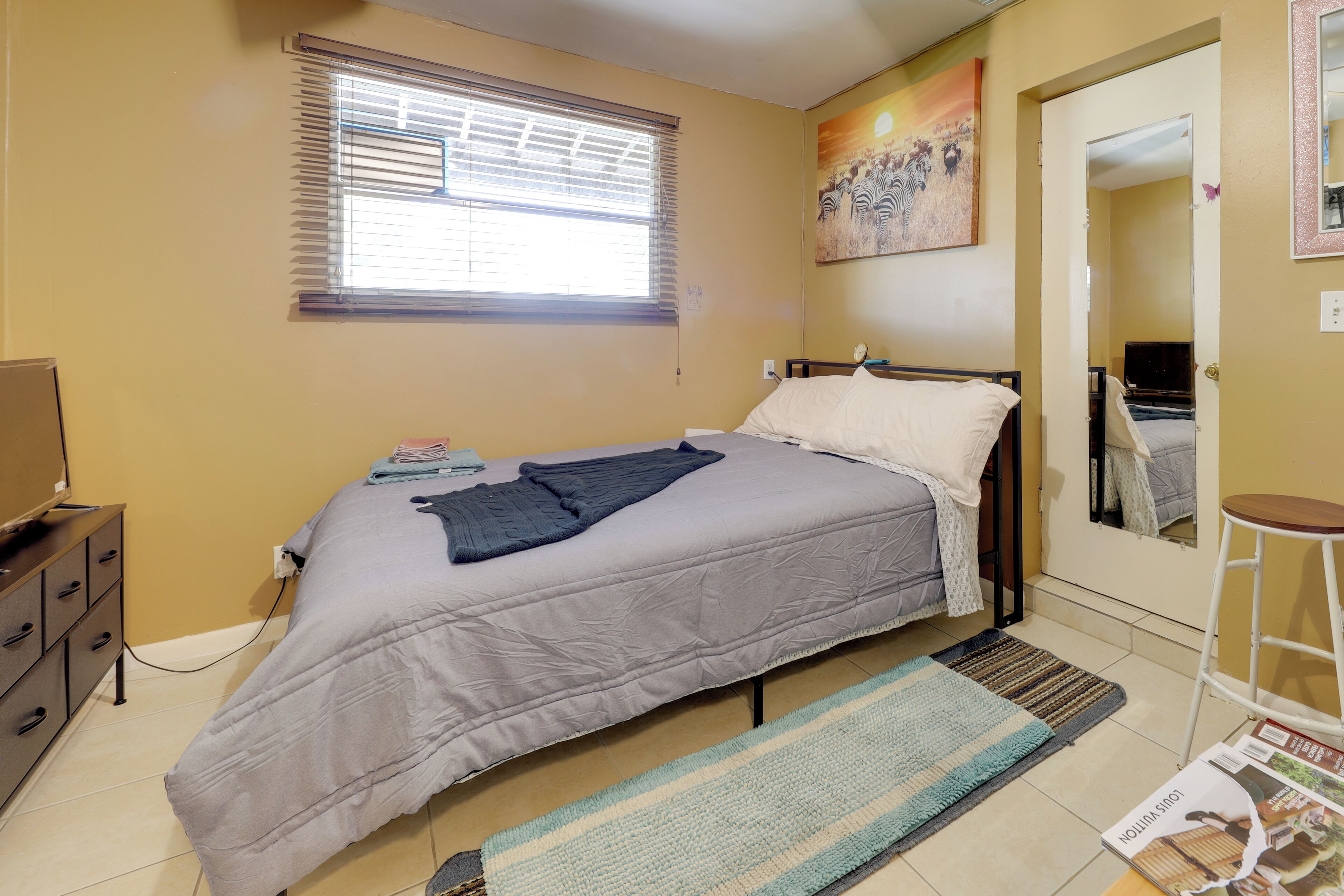 Une chambre à coucher comprenant un lit, un bureau, une chaise, une fenêtre avec des stores, un tableau accroché au mur et une porte donnant sur une autre pièce.