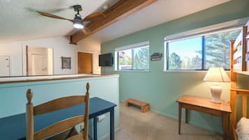 Una habitación con ventilador de techo de madera, una mesa de madera con lámpara, un banco y una ventana con vistas a árboles.