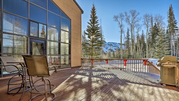 Une terrasse en bois avec une paroi en verre, des chaises d’extérieur et une vue sur les arbres et les montagnes.