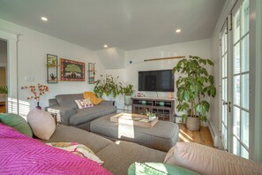 A modern living room with a grey sectional sofa, a flat-screen TV, and various plants. - Walk to Beach: Dog-friendly Ventnor City Retreat! (Ventnor City)