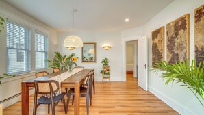 A dining area with a wooden table, chairs, and a lace table runner. There are potted plants and wall art.