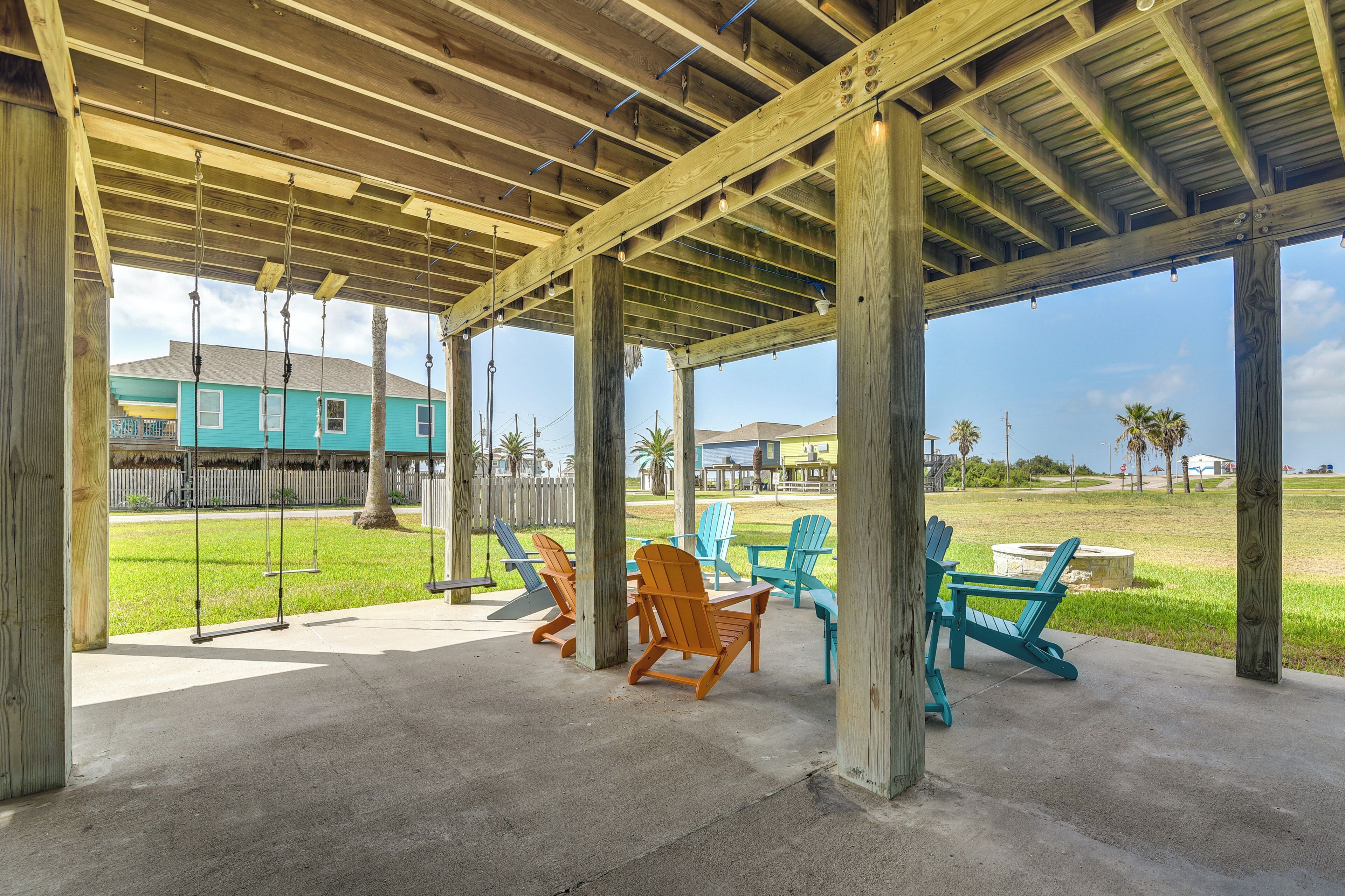 An outdoor covered patio with wooden beams and concrete flooring, featuring several Adirondack chairs and a swing set.