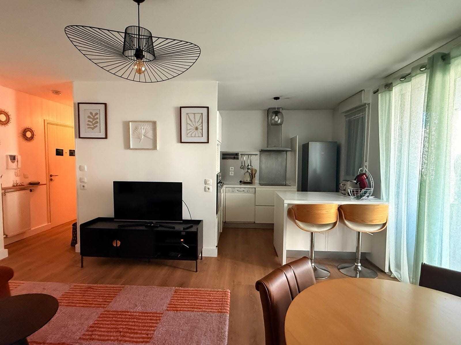 Un salon moderne doté d’un téléviseur à écran plat, d’un coin repas avec deux chaises, d’une cuisine équipée d’un réfrigérateur et d’une table à manger.