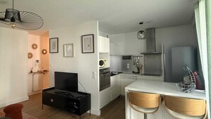 Une cuisine moderne avec des armoires blanches, une cuisinière noire et un four à micro-ondes. On y trouve un coin repas avec une table blanche et deux chaises en bois. Un meuble noir contenant un téléviseur est adossé au mur.