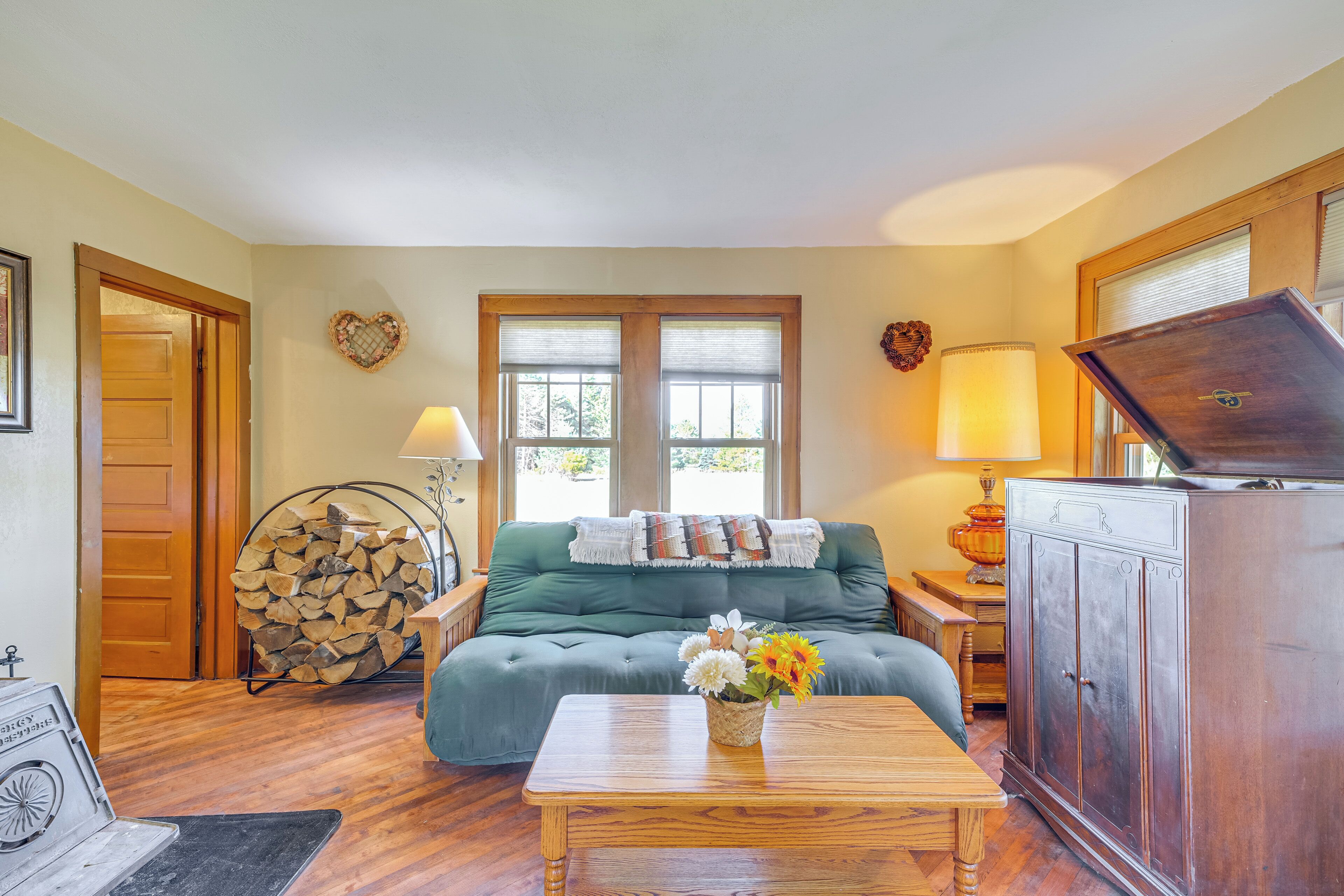 A cozy living room with a green sofa, a wooden coffee table with flowers, a stack of firewood, and a wooden cabinet.