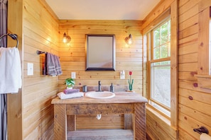 Een houten badkamer met een houten wastafelmeubel, twee wastafels, een spiegel en twee wandlampen.