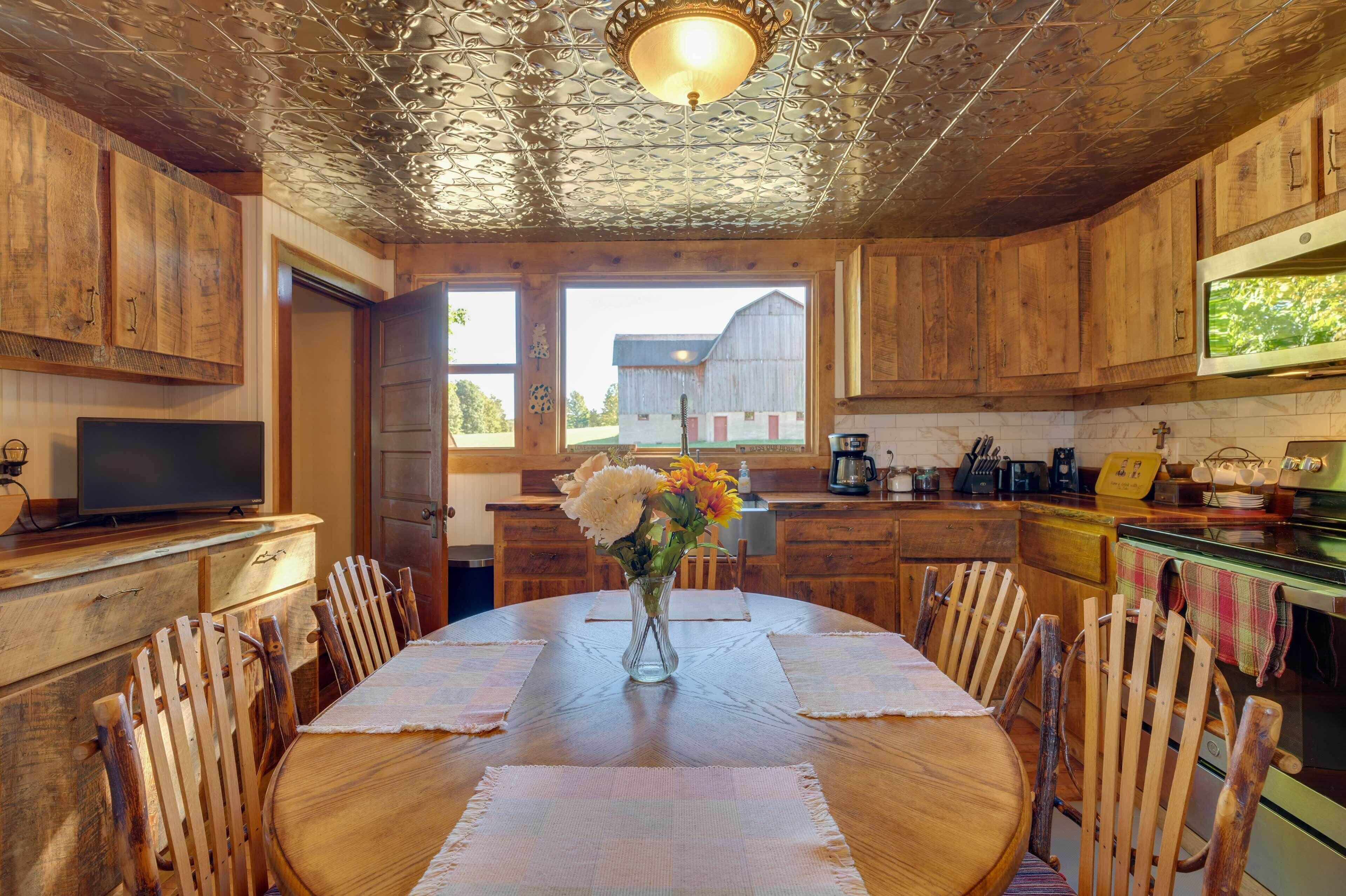 A rustic kitchen with wooden cabinets, a dining table with chairs, a vase with flowers, and a large window with a view of a barn.