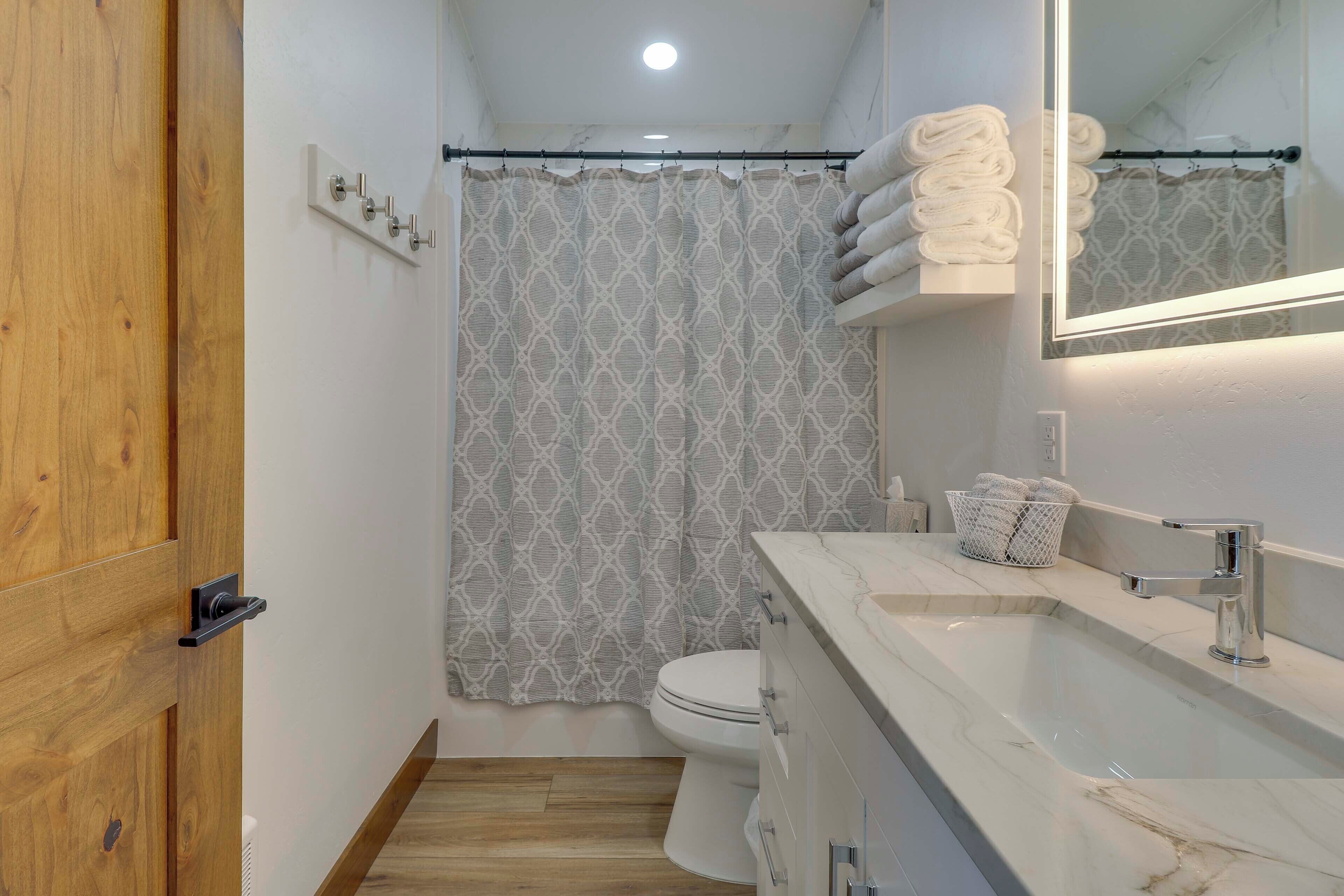A bathroom with a wooden door, a shower curtain, a toilet, a sink with a marble countertop, and a mirror.