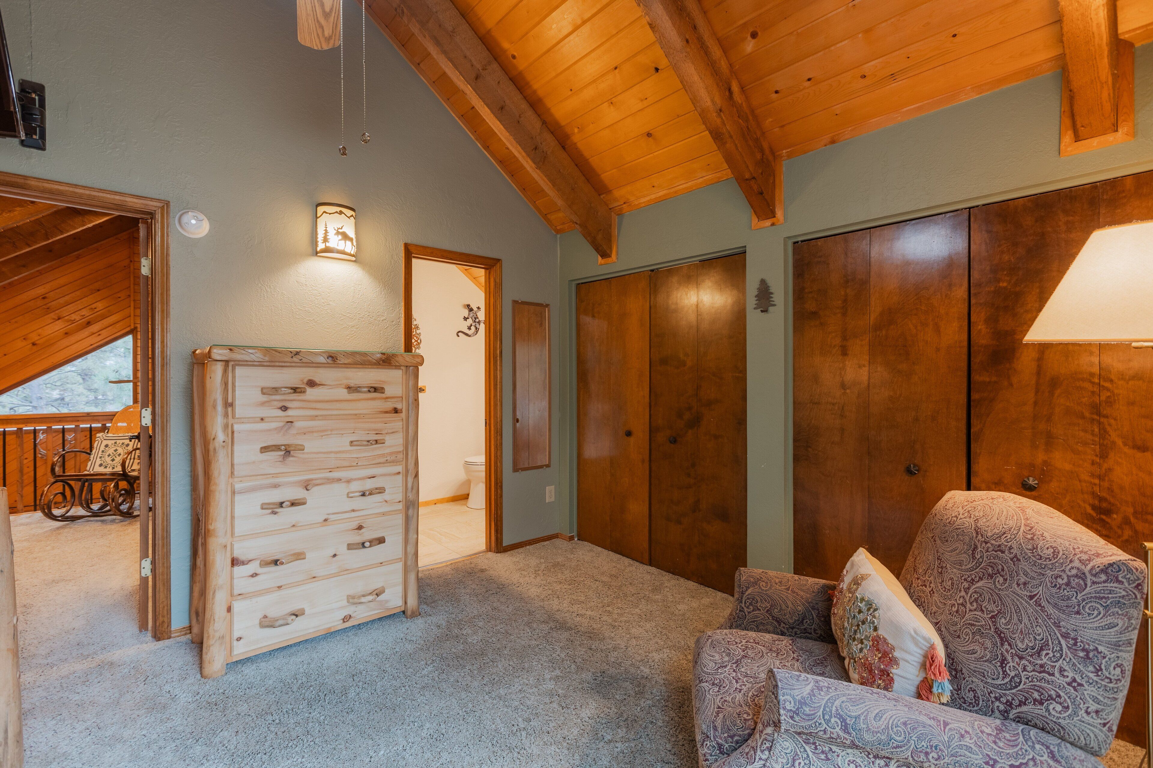 A room with a wooden ceiling, a large wardrobe, a patterned armchair, and a small table with a lamp.