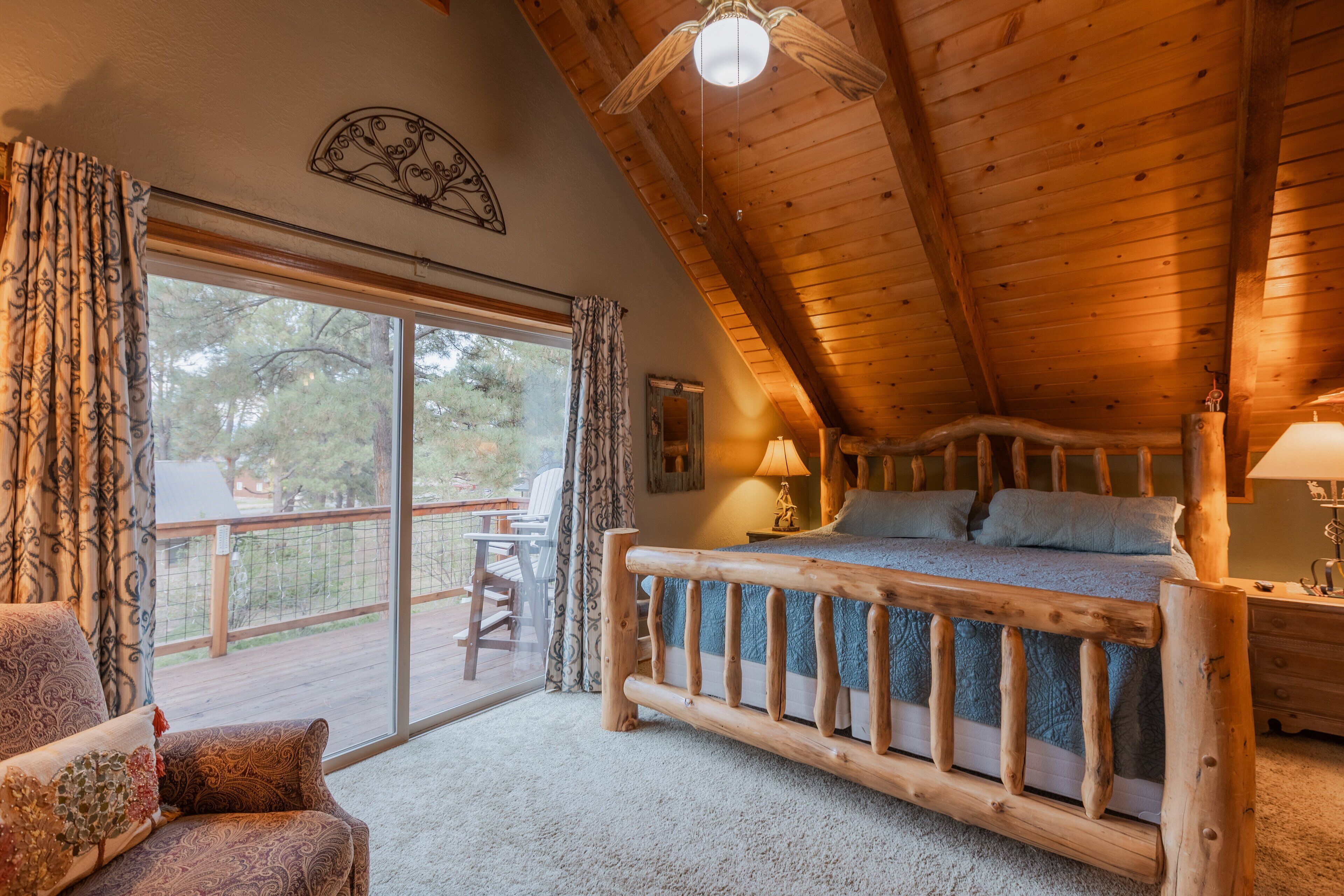 A bedroom with a wooden ceiling, a bed with blue bedding, a nightstand, a lamp, and a view of the outdoors through a sliding door.