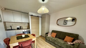 Een ruimtelijk ingerichte leefruimte met een houten eettafel, rode stoelen, een groene bank met decoratieve kussens, een keuken met witte kasten en een hanglamp.