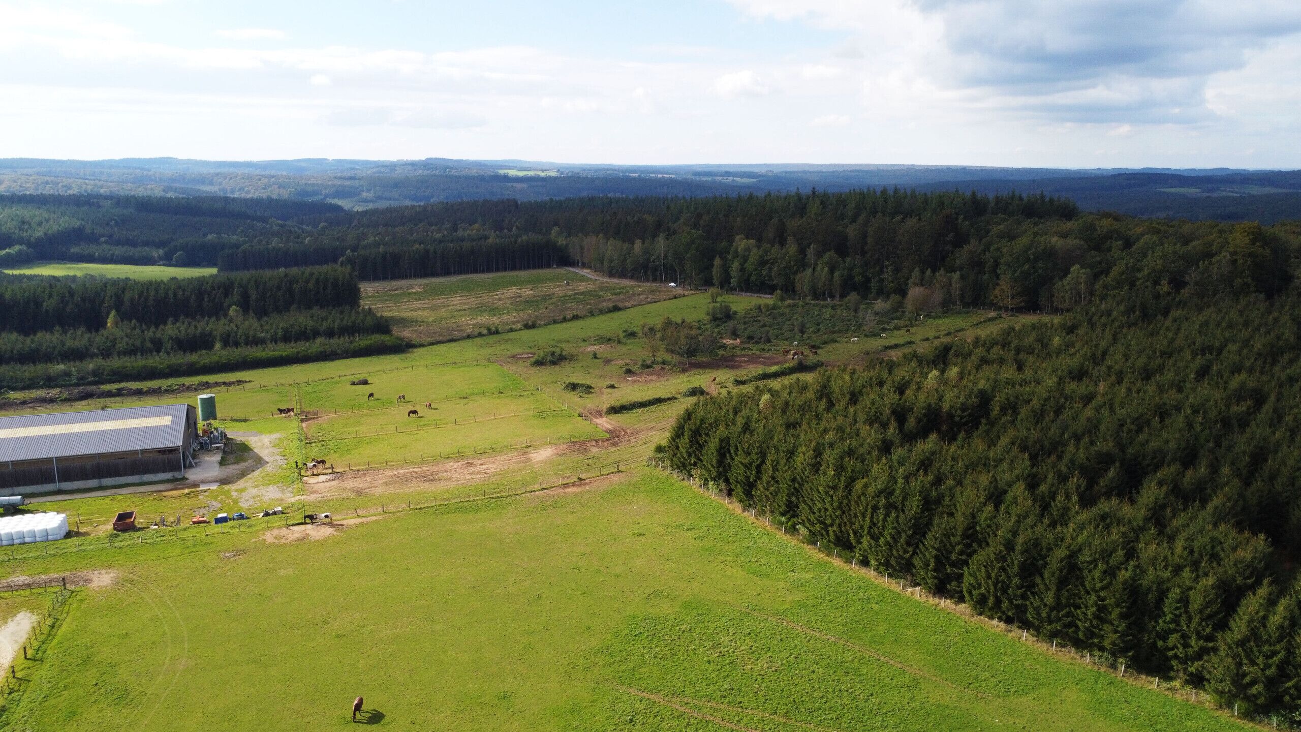 A large, open field with a building and a fenced area, surrounded by dense forest and rolling hills.