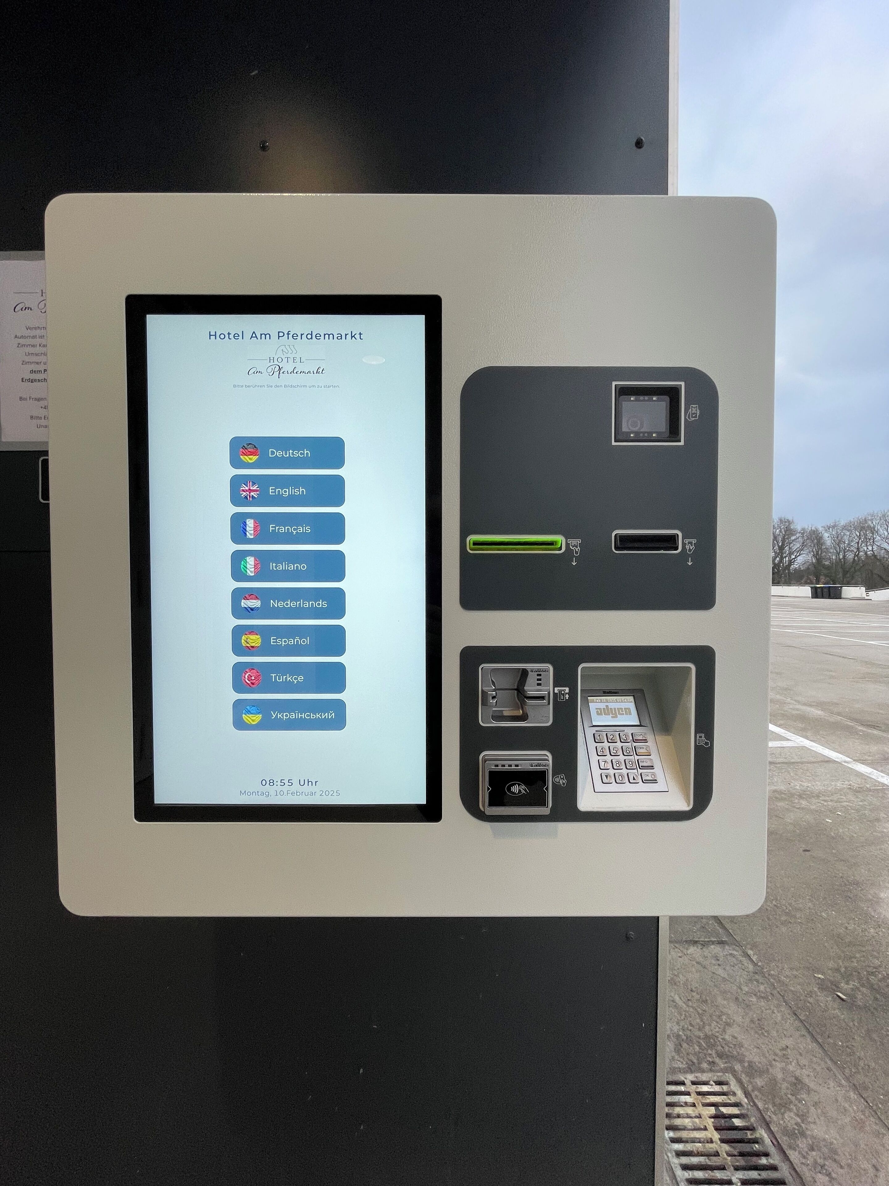 A digital kiosk with a screen displaying a hotel booking interface in multiple languages, a card reader, and a keypad.