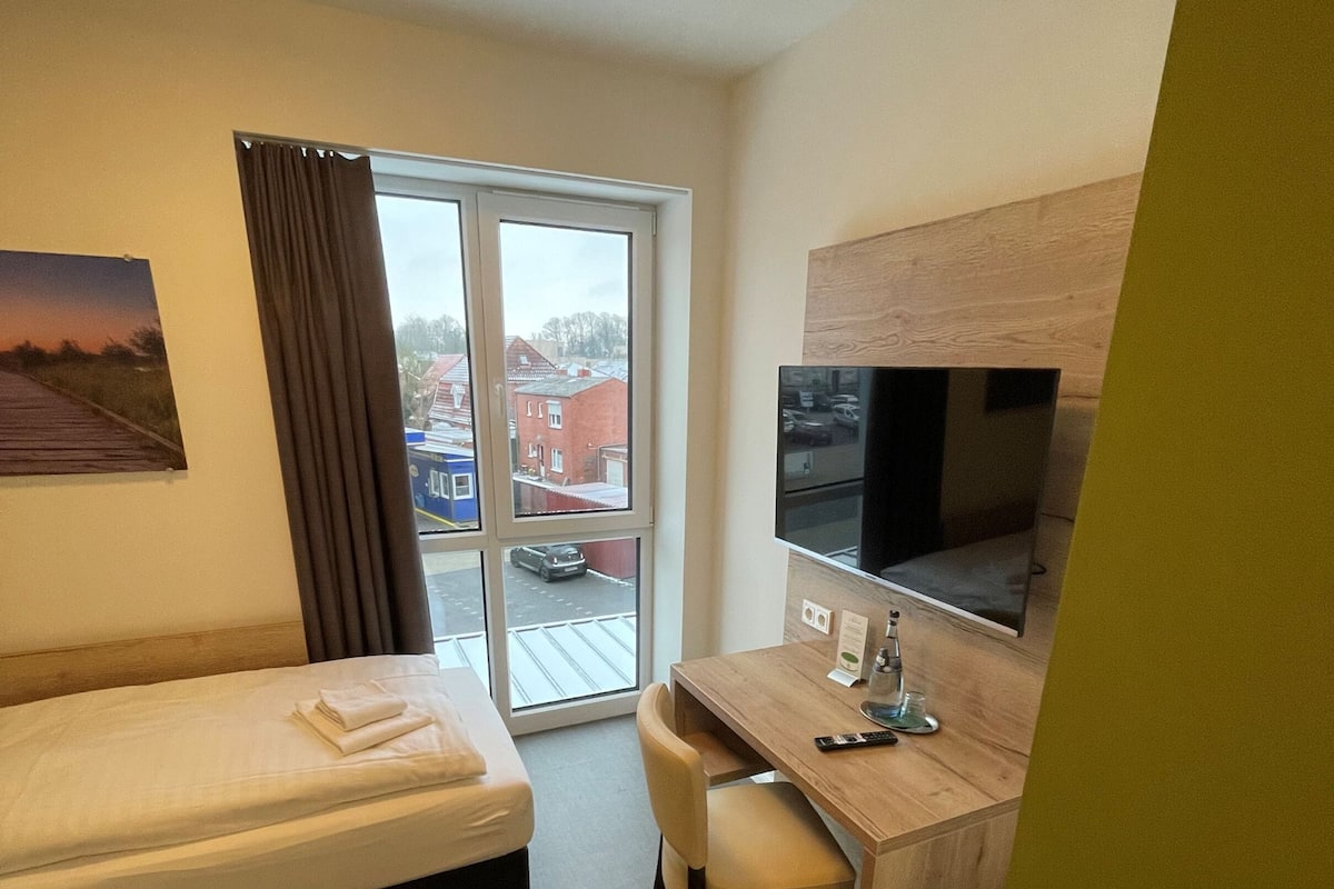 A hotel room with a bed, a desk, a chair, a TV, and a window with a view of buildings and a car.