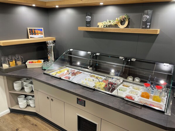 Zona de desayuno tipo buffet con diversas opciones de comida, una máquina de café y un cartel con una cita.