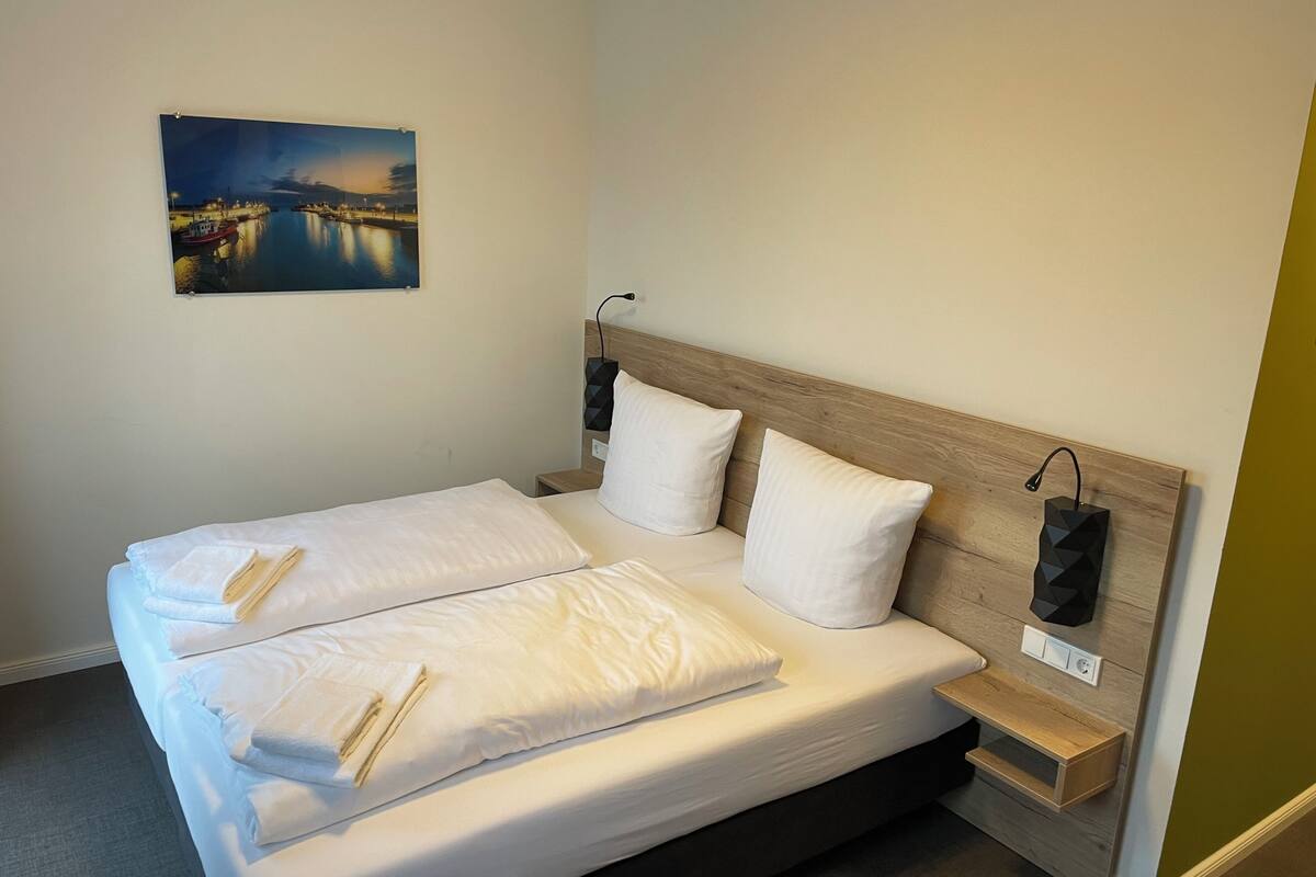 A neatly made double bed with white linens and pillows, a small bedside table with a lamp, and a wall-mounted power outlet.