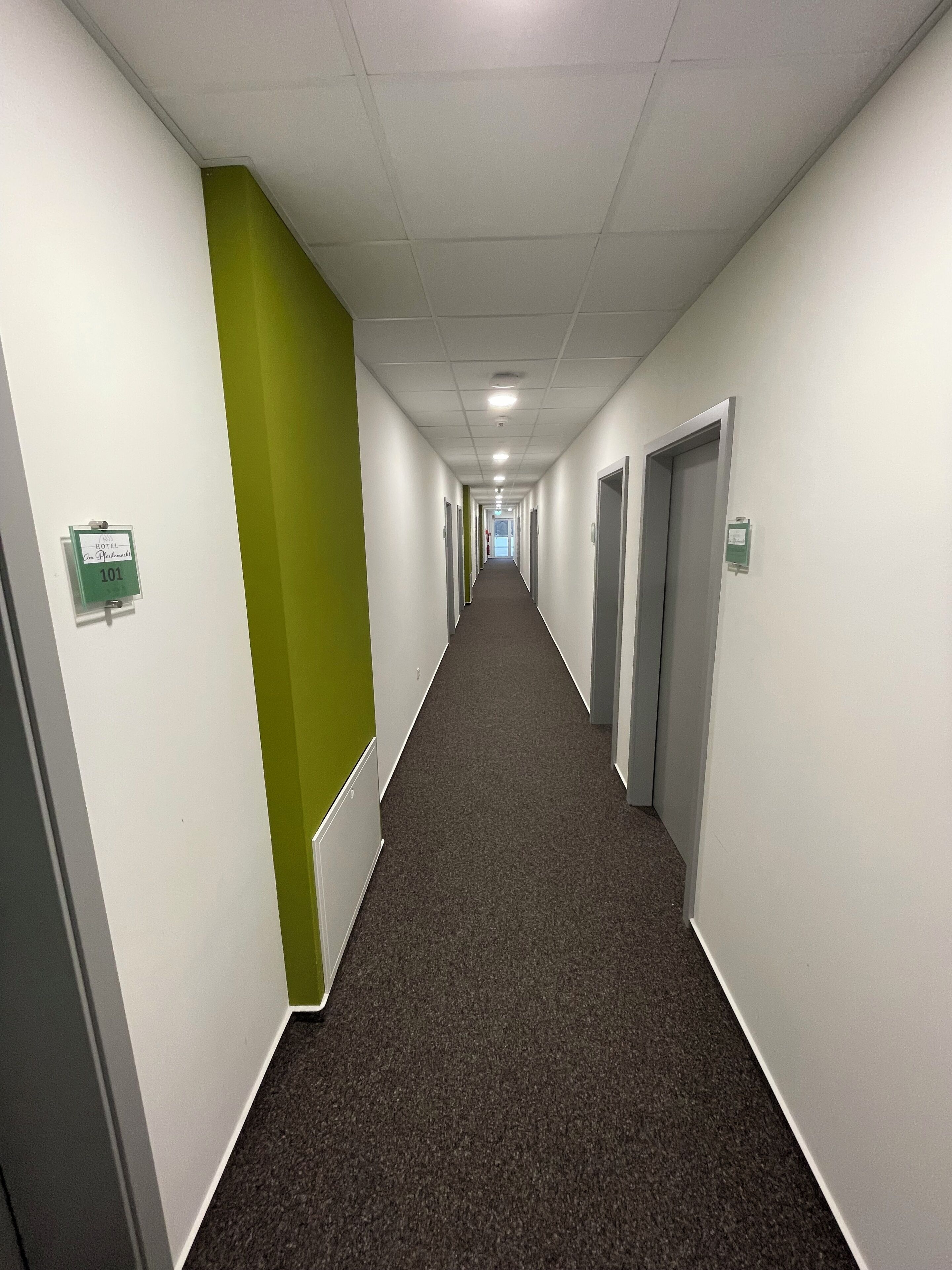 Un long couloir étroit aux murs blancs, avec un mur d’accent vert sur le côté gauche. Le sol est recouvert de moquette et il y a des portes de part et d’autre du couloir.