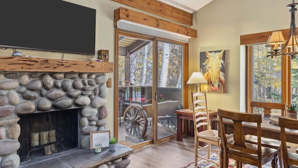 A stone fireplace with a television above it, wooden beams on the ceiling, and a dining area with chairs and a table. - Outpost: Columbine 1024 - Aspens Location! (Wilson)