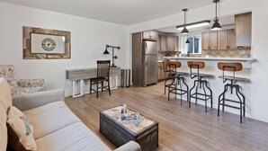 A modern living room with a sofa, a coffee table, and a kitchen area with a refrigerator, stove, and bar stools.