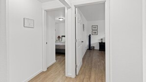 A hallway with white walls and wooden flooring, leading to a bedroom with a bed and a nightstand.