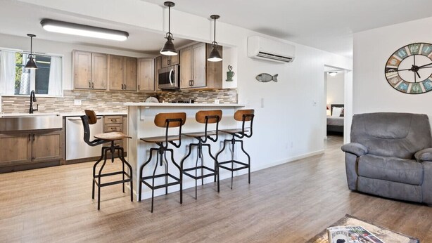 A modern kitchen with wooden cabinets, a central island with bar stools, and a grey sofa in the living area.