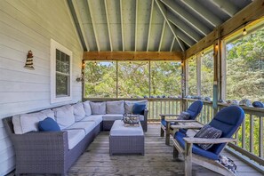 A covered porch with a cushioned seating area, a small table, and a view of the surrounding greenery. - Gray Whale: Fenced Yard | Screened Porch (Ocracoke)