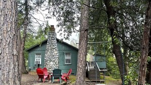 A rustic cabin with a stone chimney, surrounded by trees. There are two red Adirondack chairs and a green Adirondack chair outside. A wooden staircase leads to a deck.
