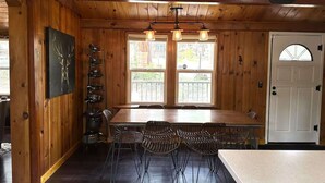 A wooden interior with a dining table and chairs, a kitchen area with pots and a hanging rack, and a door with a window.