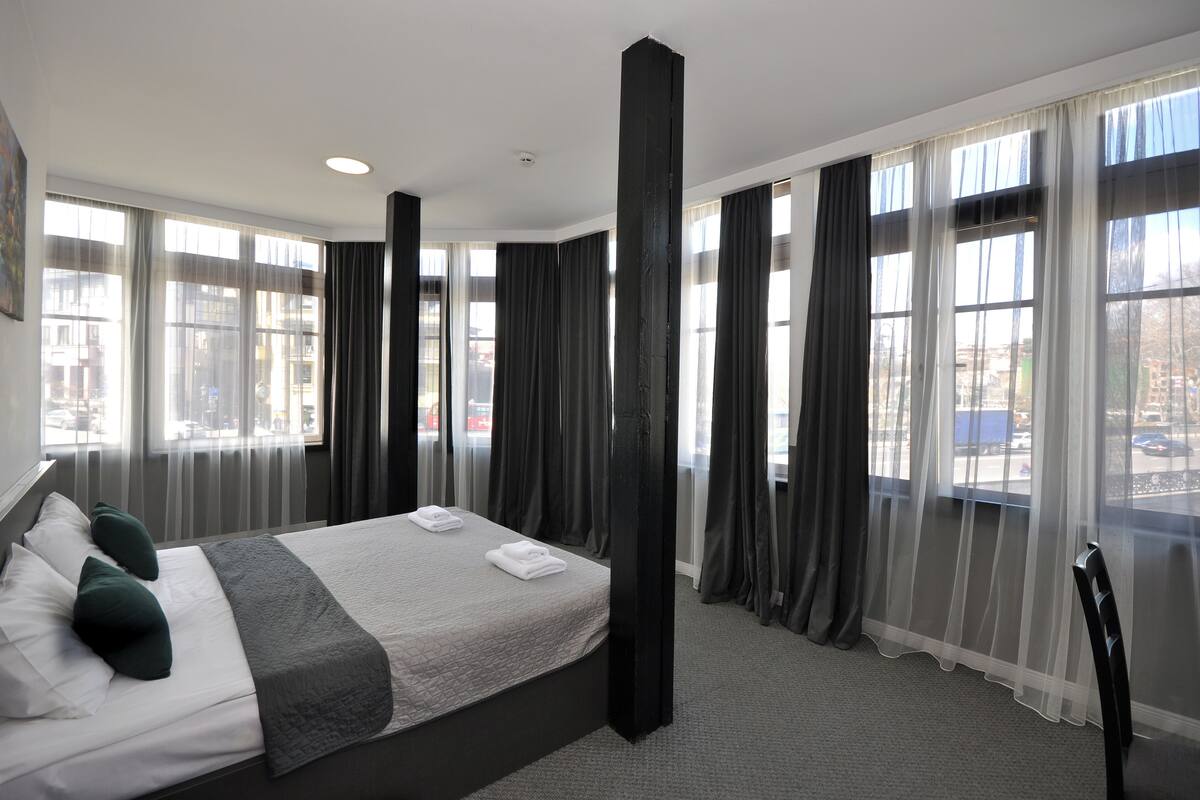 A hotel room with a large bed, a chair, and a view of the street through sheer curtains.