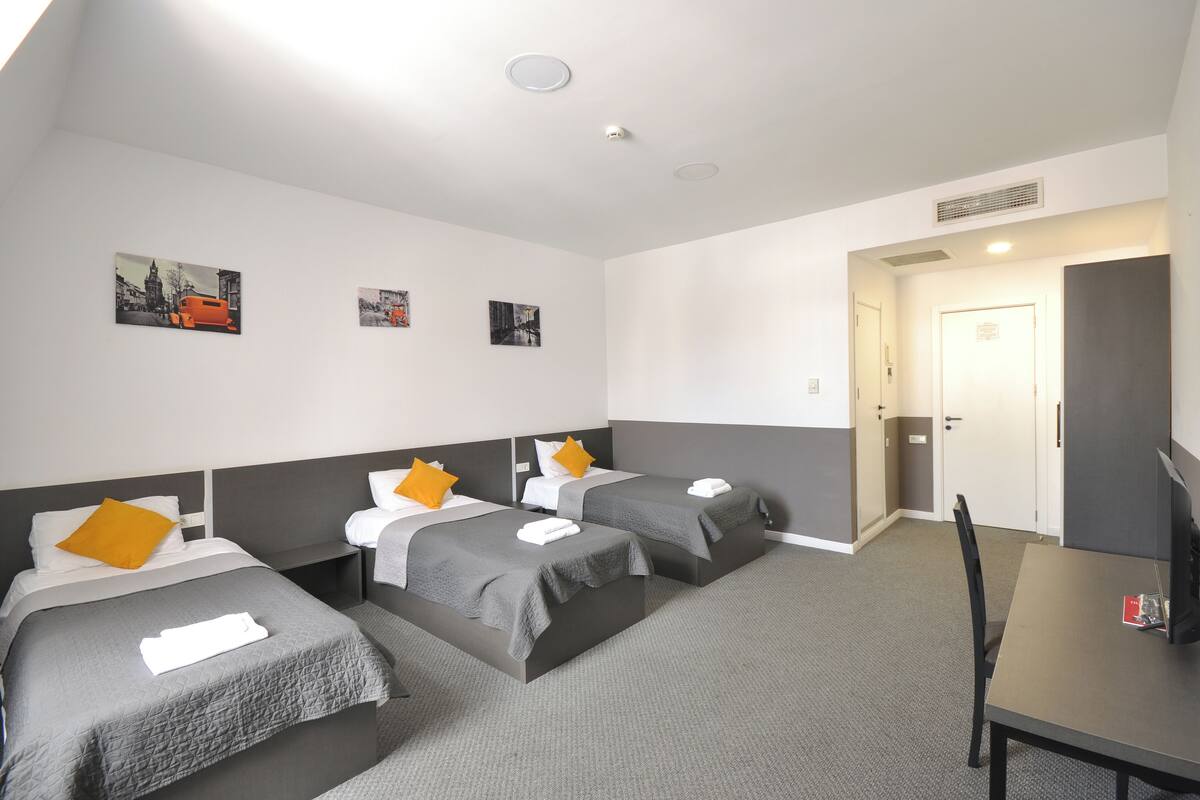 A hotel room with two beds, a desk, and a TV. There are pictures on the wall and a door to the right.