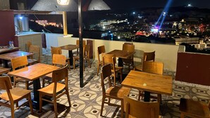 Area pranzo all'aperto con tavoli e sedie in legno, pavimento decorato e vista sulla città illuminata di notte.