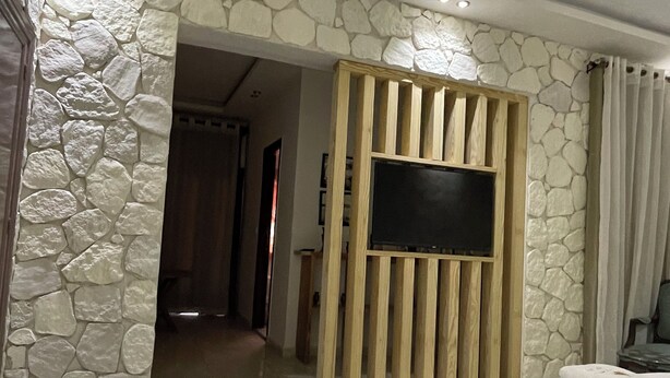 Camera d'albergo con due letti, pareti in pietra, un divisorio in legno con un televisore e un bagno con doccia e toilette.