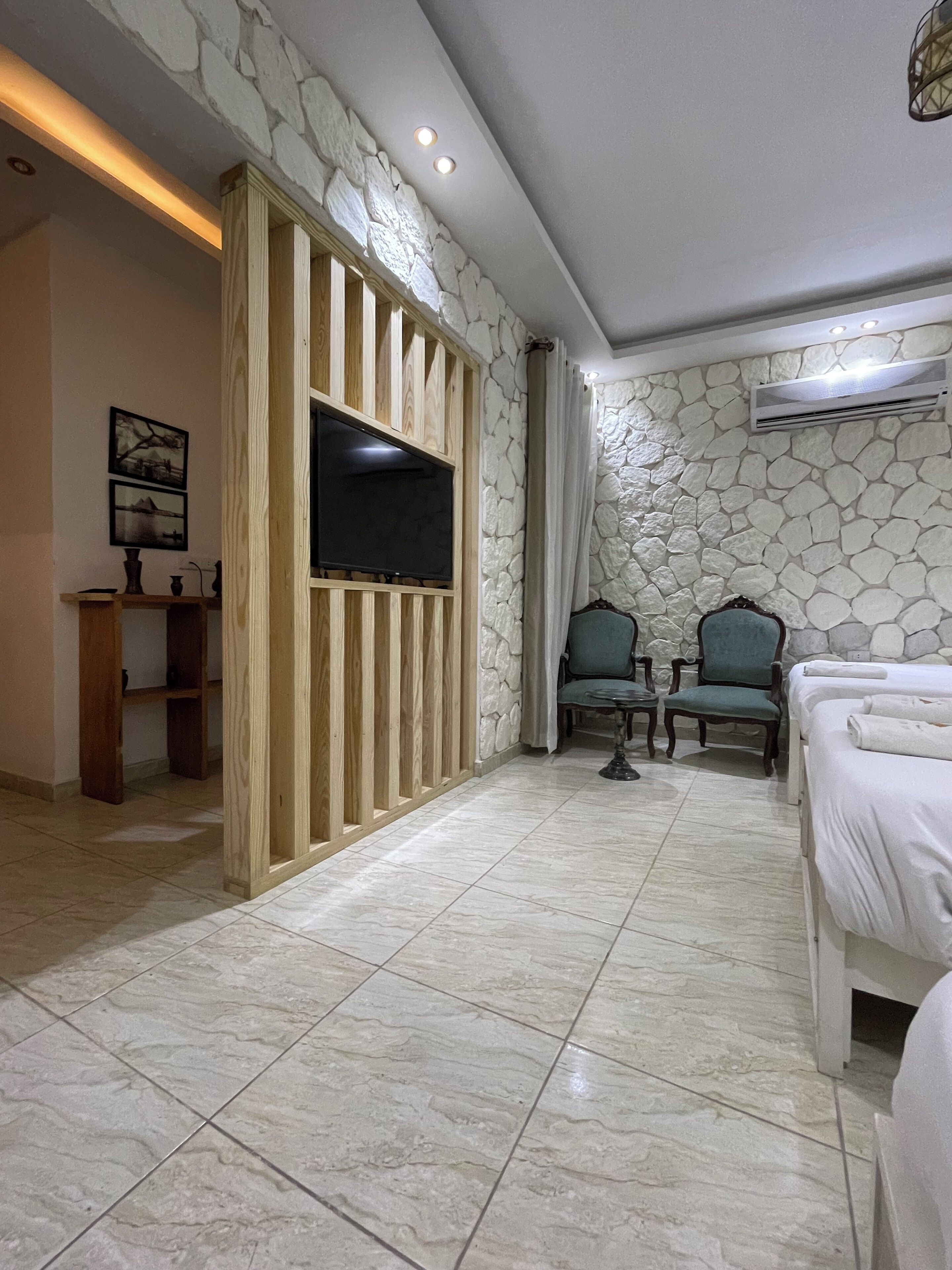 A hotel room with a stone wall, a flat-screen TV mounted on a wooden panel, two chairs, a bed, and a small table.