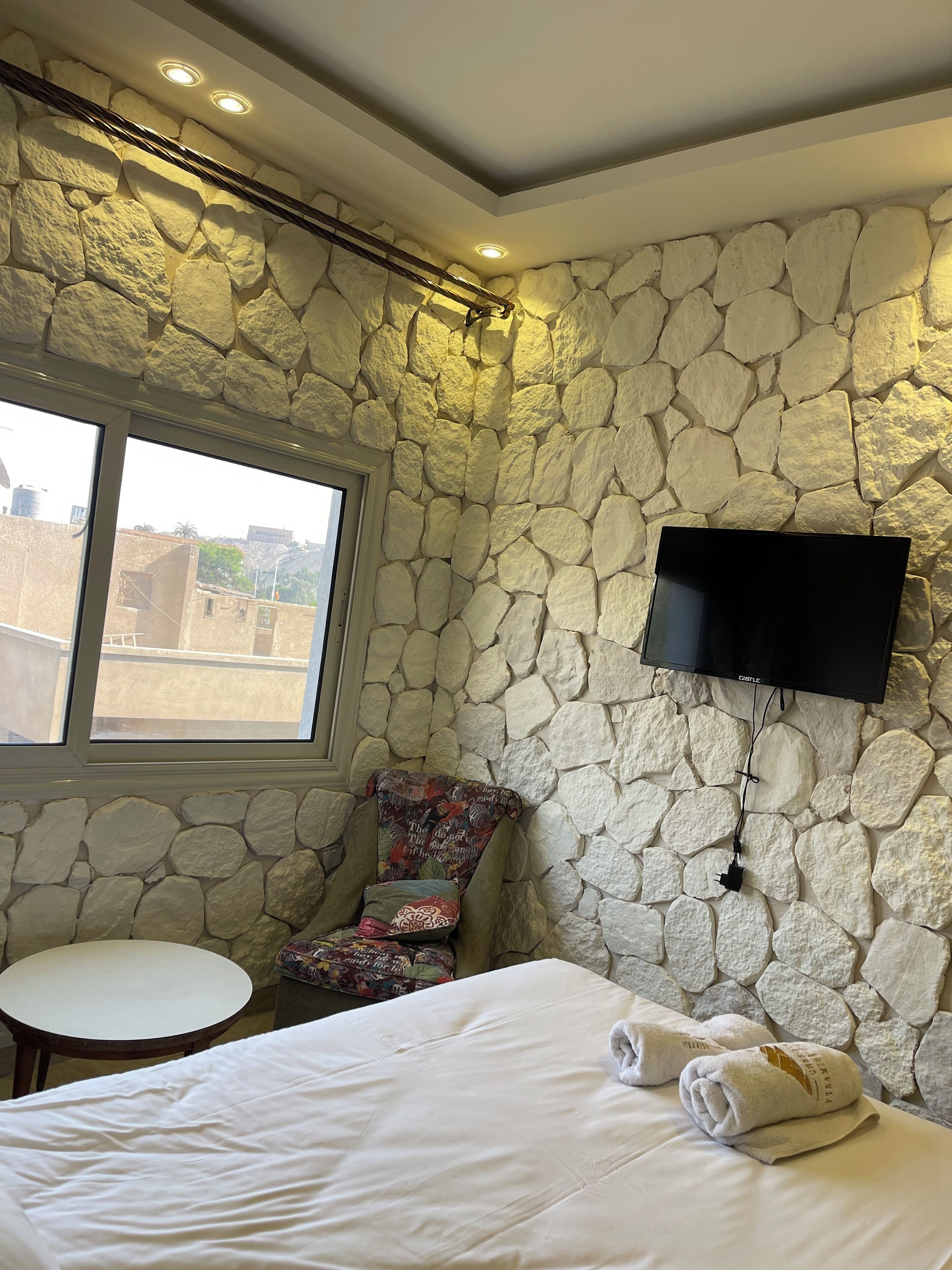 A hotel room with a stone wall, a bed with white linens, a flat-screen TV mounted on the wall, a small round table, and a colorful chair.