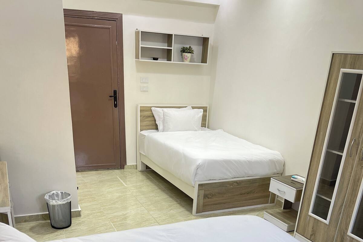 Habitación de hotel con dos camas, una mesita de noche pequeña, un cubo de basura, un armario y un estante con una planta.