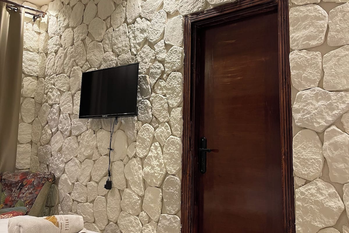 Un dormitorio con pared de piedra, puerta de madera, televisor de pantalla plana montado en la pared, cama con colcha blanca y una toalla sobre la cama.