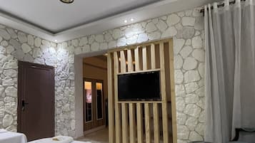 Une chambre d’hôtel avec deux lits, un téléviseur fixé sur un panneau en bois, des éléments de décoration en pierre au mur et un sol en pierre.