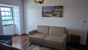 A room with a beige sofa, a patterned rug, a painting of a house, and a window with a view of the sea. - St. John's House (São João)