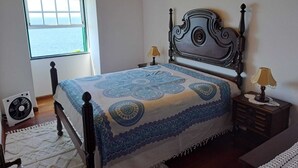 A bedroom with a large bed, a window with a view of the sea, a bedside table with a lamp, and a fan on the floor. - St. John's House (São João)