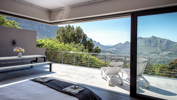 A modern bedroom with a large bed, a balcony with a view of mountains, and a dining area with a table and chairs. - Stylish apartment with Endless Views & Privacy (Cape Town)