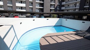 Ein Pool auf dem Dach mit Holzterrasse und Blick auf ein mehrgeschossiges Gebäude im Hintergrund.