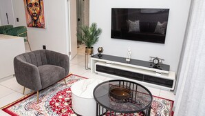 Ein modernes Wohnzimmer mit einem grauen Sessel, einem runden Glastisch, einem weißen Hocker, einem roten und weißen Teppich, einem weißen Konsolentisch mit Globus und Uhr sowie einem großen Fernseher an der Wand.