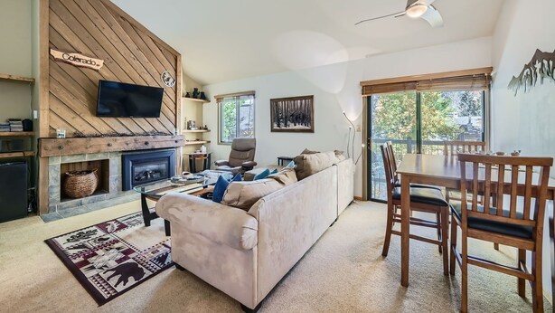 A living room with a fireplace, a television mounted on a wooden wall, a sofa, a dining table with chairs, and a view through a large window. - 1147 Flying Dutchman 1 Bedroom Condo (Keystone)