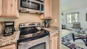 A kitchen with a stainless steel microwave, a coffee maker, and a stove. There is a window with a view of trees and a chair in the adjacent room. - 1147 Flying Dutchman 1 Bedroom Condo (Keystone)