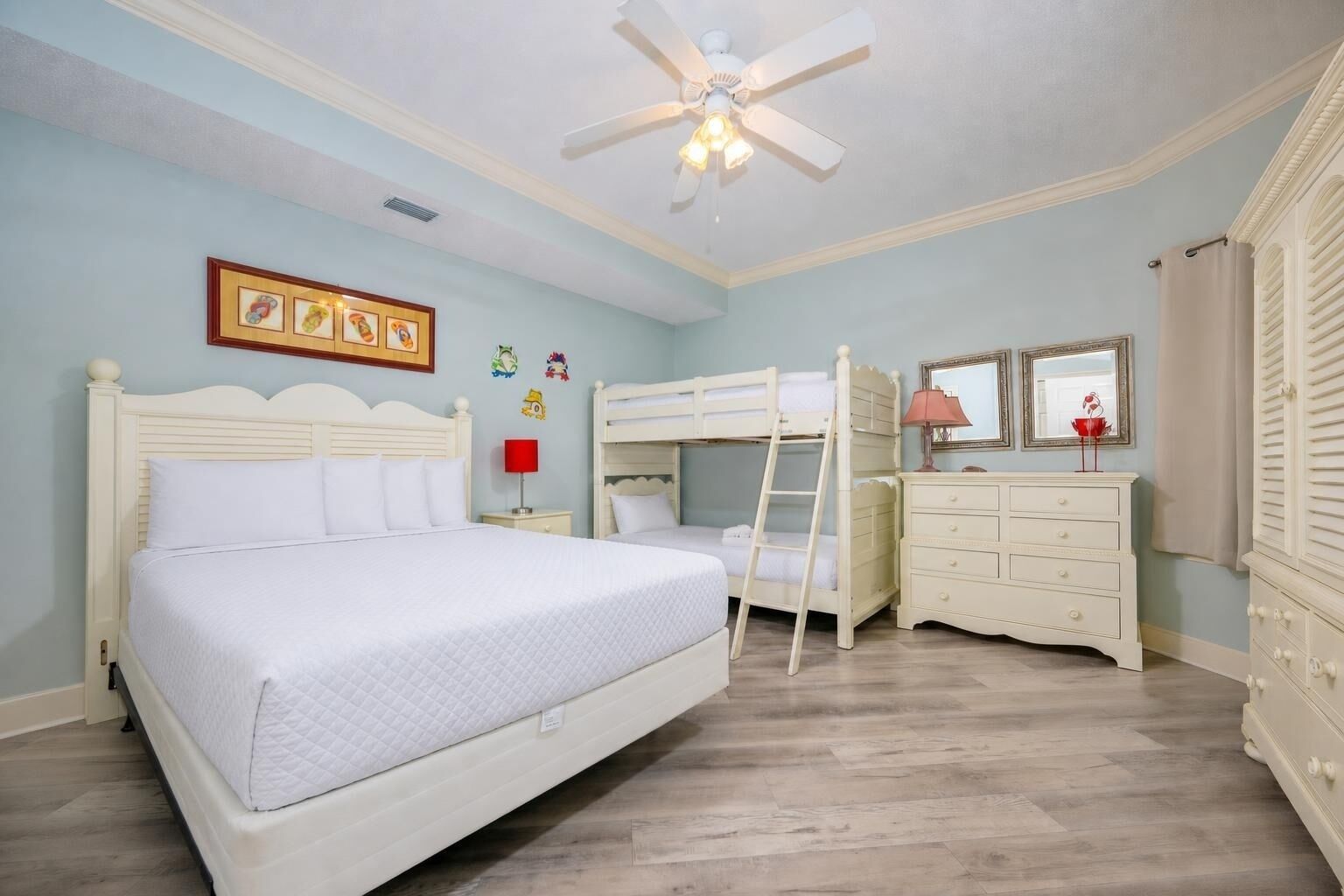 A bedroom with a bunk bed, a single bed, a dresser, a vanity, a ceiling fan, and a framed picture on the wall.