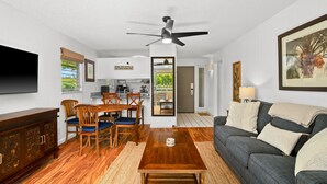 A modern living room with a dark grey sofa, wooden dining table, and chairs. A ceiling fan is visible, and a flat-screen TV is mounted on the wall. - Kauai Plantation Hale Suites E12 1 Bedroom Condo (Kapaa)