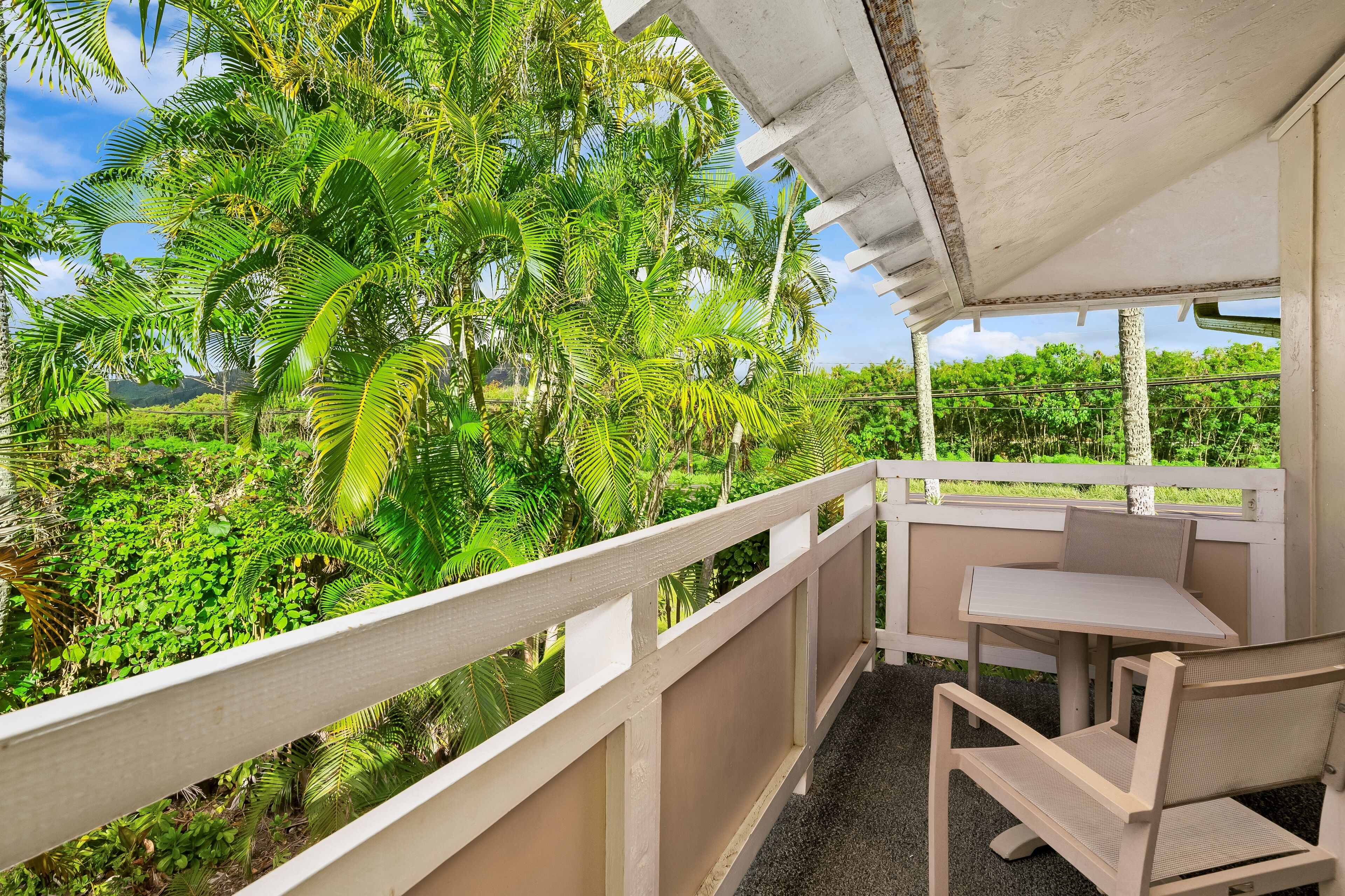 A balcony with a white railing, a small table, and a chair, overlooking a lush garden with various plants and trees.