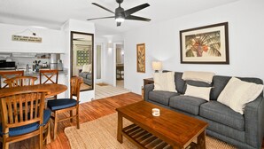 A living room with a grey sofa, wooden coffee table, and a dining area with a wooden table and chairs. - Kauai Plantation Hale Suites E12 1 Bedroom Condo (Kapaa)